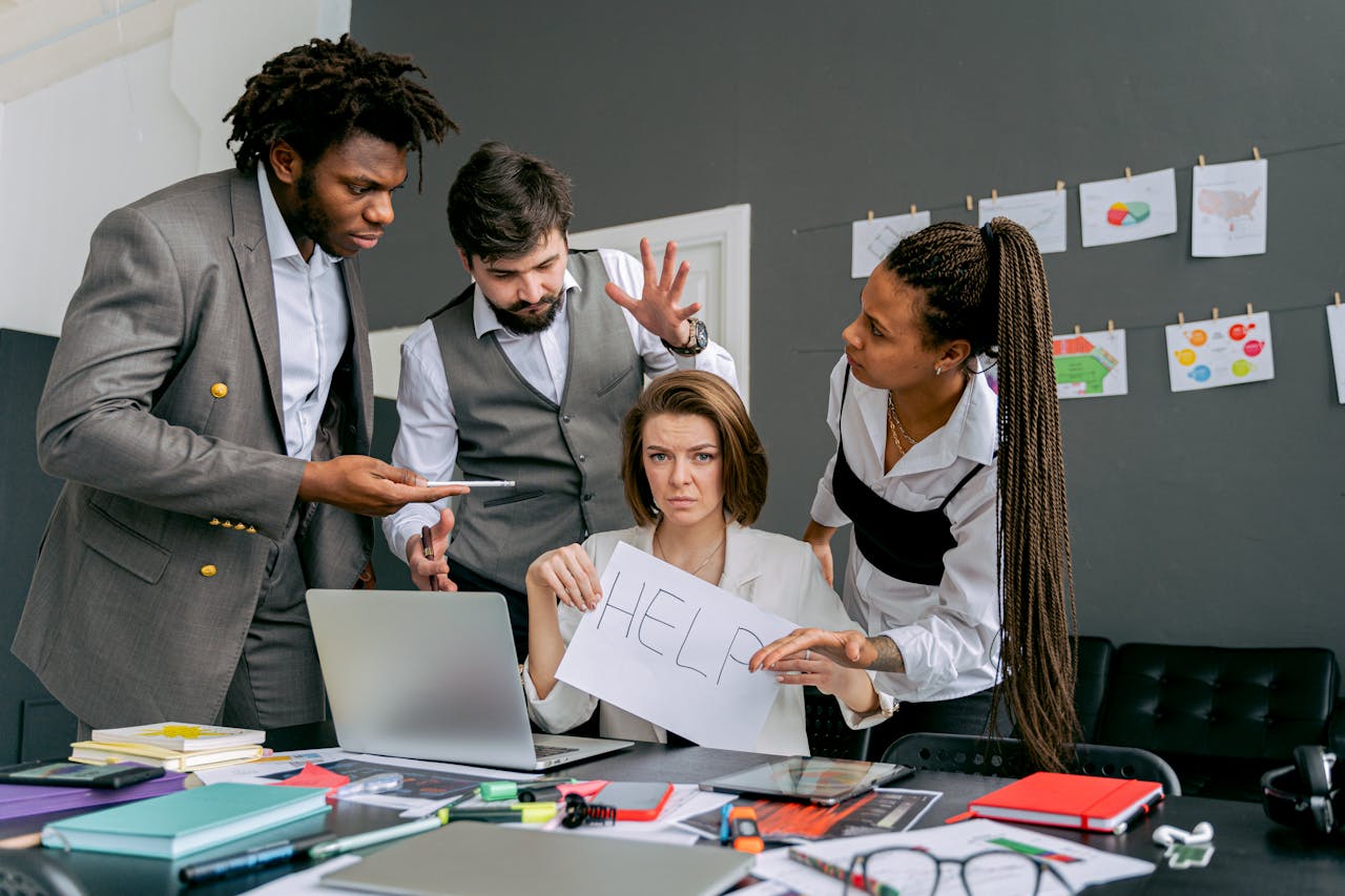 Office scene with diverse colleagues around a stressed woman holding a sign that says HELP. Focus on teamwork dynamics.